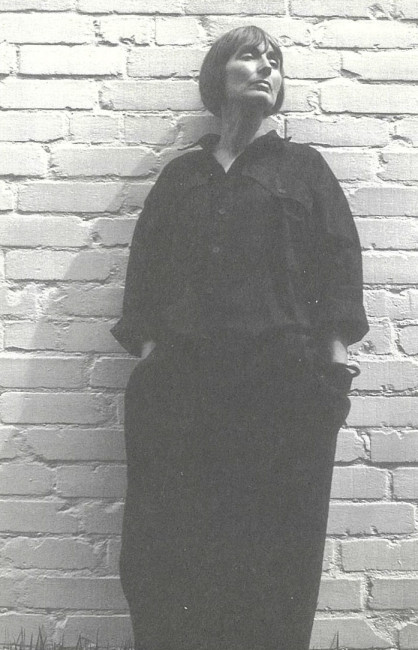 Eine in schwarz gekleidete ältere Dame lehnt an einer weißen Ziegelwand